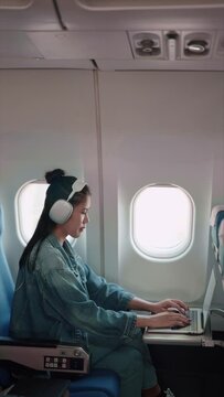 Asian female traveler working on her laptop and wearing headphones while flying, getting things done during the plane ride for a productive holiday trip