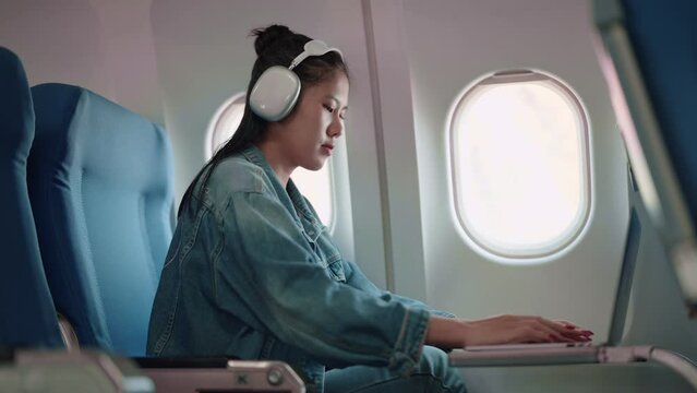 Asian female traveler working on her laptop and wearing headphones while flying, getting things done during the plane ride for a productive holiday trip