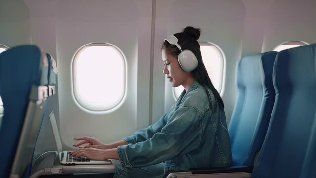 Asian female traveler working on her laptop and wearing headphones while flying, getting things done during the plane ride for a productive holiday trip