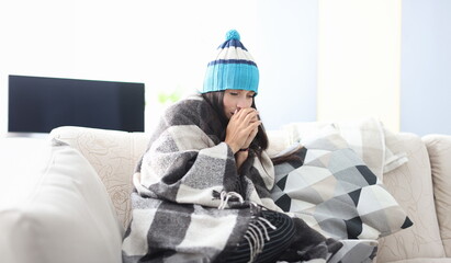 Sick woman in hat sit on sofa covered with blanket. Person hold hands together near his mouth and...