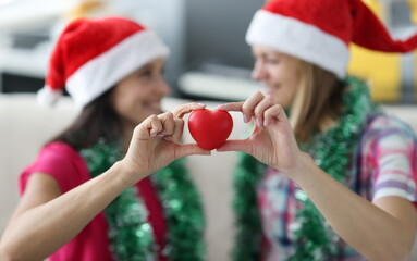 Red shape heart close up. Two woman in santa claus hat and tinsel hold symbol of love in hand.