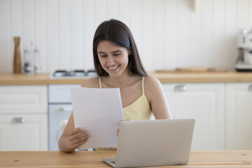 Young positive woman sits at table with laptop in kitchen holding paper notice, enjoy pleasant...