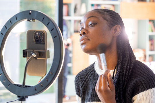 This image depicts a social media influencer woman engaged in her beauty routine, applying makeup with a brush in front of a ring light mirror that illuminates her face. Her expression is one of