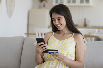 Smiling young woman enjoy electronic shopping at home, sit on sofa hold credit card and smartphone, making remote order, buying clothes, goods and gifts through e-commerce application. Secure payment