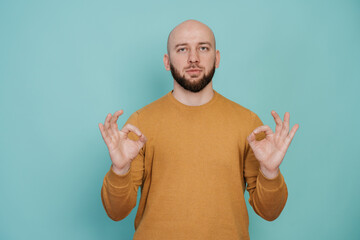 Fototapeta premium calm bald bearded man in casual holds hands in meditative gesture against turquoise background. focus concept. Mental balance concept. Keep calm.