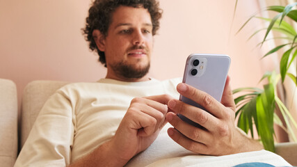 Young man with curly hair and beard, sitting on sofa in cozy living room, using mobile phone