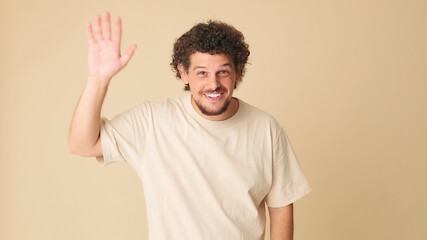 Smiling guy with curly hair dressed in beige t-shirt looking at the camera and waving his hand in welcoming gesture, saying hello to someone, isolated on beige background in the studio