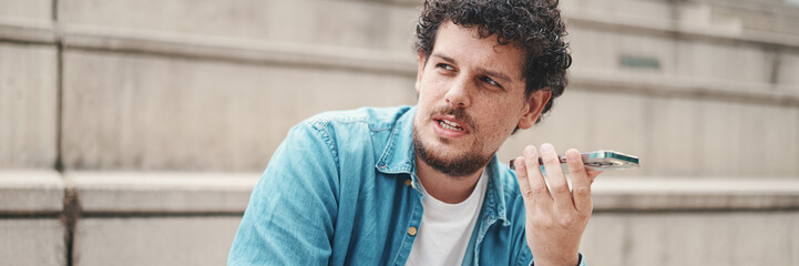 Fototapeta premium young bearded man in denim shirt sitting on high steps and using mobile phone. man sends voice message to smartphone