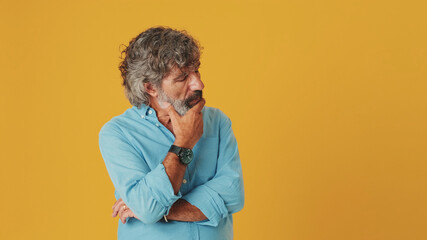 Elderly grey-haired bearded man wears blue shirt, thinks rubs chin comes up with ideas, isolated on orange background in studio