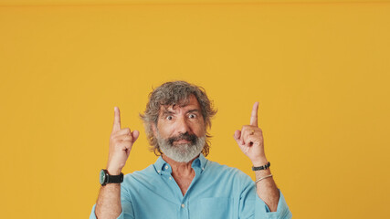 Elderly grey-haired bearded man wears blue shirt looking up points fingers up at at the copy space...