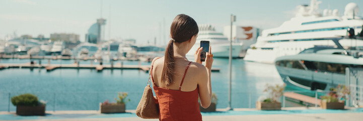 Cute woman takes photos, videos of the seaport on a mobile phone. girl uses social networks and...