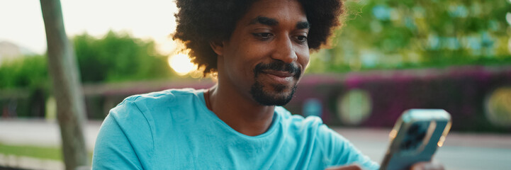 Close-up of young African American man in blue t-shirt sitting on park bench and talking on...