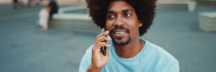 Closeup young African American man in light blue t-shirt sitting on city park bench and talking on...