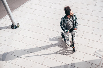 Trendy smiling bearded man in casual clothes riding electric scooter on a road in urban background. Handsome model posing in the street. Hipster guy with curly hairstyle. Top view