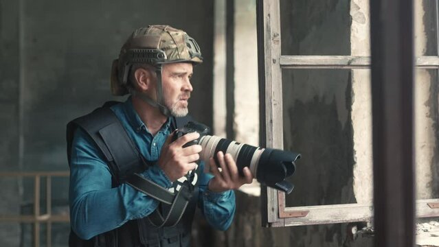 Handsome older Caucasian male peeking rapidly into window from behind wall of abandoned building to make photo. Working in dangerous environment. Wearing helmet and vest, Using professional gear.