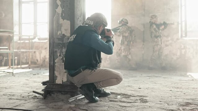Rear view of reporter taking photo on camera of military soldiers hiding from shelling in abandoned building. Military concept. People wearing safety helmets and bulletproof vests during confrontation