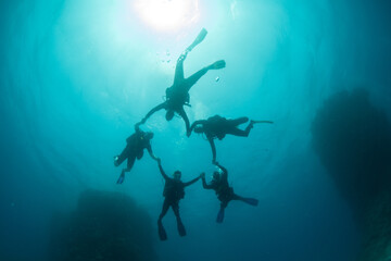 Silhouettes of a group of scuba divers forming a circle holding hands around the sun underwater