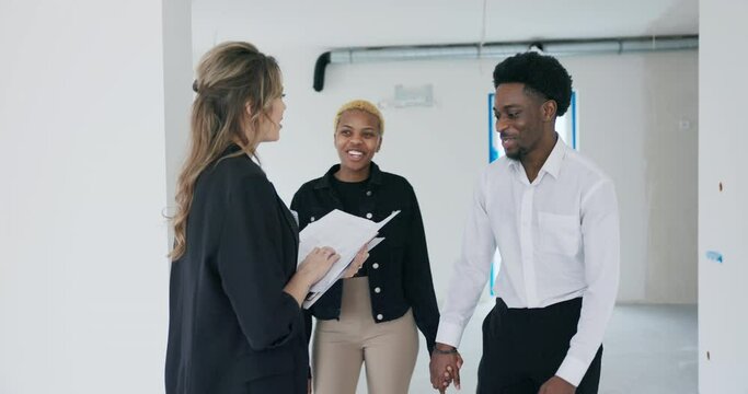 Blonde real estate agent stands, contract and house plan in hand, eagerly awaiting the arrival of the African American couple. As they enter exchange warm waves with the agent, signaling enthusiasm.