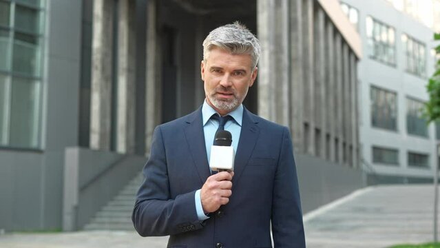 Attractive Caucasian reporter speaking to camera while holding microphone. Intelligent man actively telling latest events and interesting news of city. Dissemination of information in mass media.