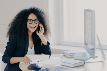 Joyful businesswoman on phone call at desk with computer and notebooks