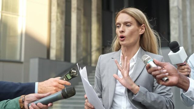 Crowd of correspondents waiting for interview with famous successful Caucasian woman who leaving her office. Concentrated political female with documents avoids talking to persistent journalists.