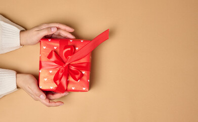 Female hands holding a gift box with a red ribbon on a brown background.