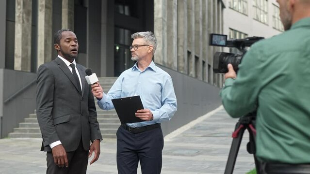 Camera view of mixed-raced people recording interview while standing near large building. Handsome politician with black suit having dialogue with diplomatic correspondent outdoor.