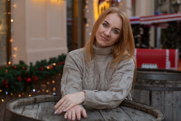 Smiling woman in sweater next to a table in a outdoor cafe. Christmas decorations in the background. Cold season, walk in the city