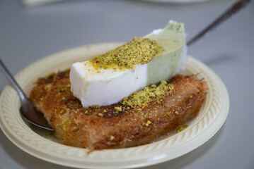 A slice of Kanafeh, a traditional middle eastern delicious dessert, served in a white plate with topping of pistachios , and vanilla and pistachio ice cream