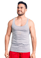 Young handsome man wearing swimwear and sleeveless t-shirt looking away to side with smile on face, natural expression. laughing confident.