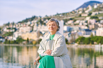 Outdoor portrait of happy mature 50 - 55 year old woman, enjoying nice spring day by the lakeside, wearing grey coat - obrazy, fototapety, plakaty