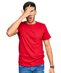 Young handsome man wearing casual red tshirt peeking in shock covering face and eyes with hand, looking through fingers with embarrassed expression.