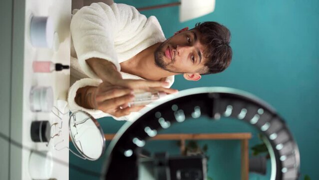 Handsome Arabic beauty blogger in bathrobe present serum for skin rejuvenation in home interior. Influencer record video review blog using smartphone