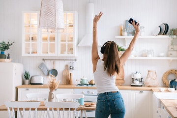rear view of a positive girl in jeans dancing in the kitchen at home using headphones and a phone that raised her hands up shows a victory sign a fun weekend at home