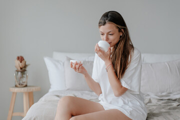 American girl with long hair is sitting on the bed in a T-shirt, holding a cream in her hands,...