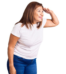 Middle age latin woman wearing casual white tshirt very happy and smiling looking far away with hand over head. searching concept.