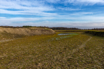 A landscape photo captures a grassy field with a small pond in the center, under a clear blue sky.