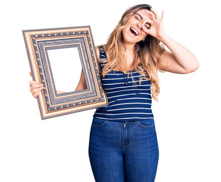 Young caucasian woman holding empty frame smiling happy doing ok sign with hand on eye looking through fingers