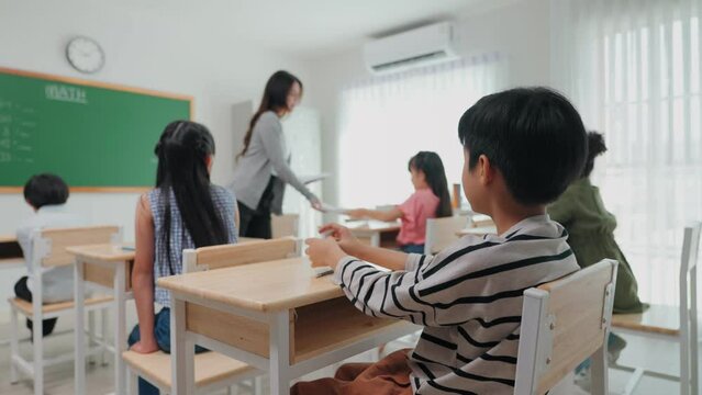 Young teacher woman handing out papers for exams to children sit on desk in classroom at school. Diverse primary students preparing final exam at elementary school. Assessment and measure education.