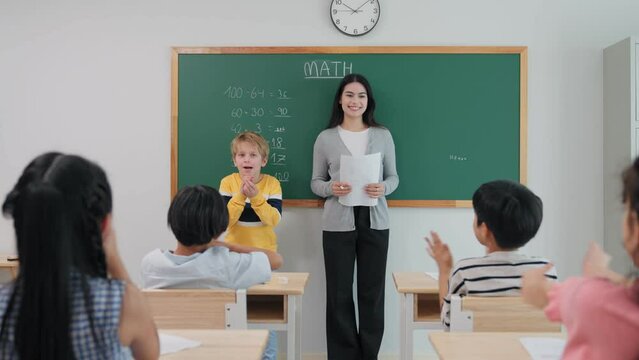 Smart caucasian cute boy writing answer math formula on blackboard with chalk during lesson at school. Young teacher woman and diverse student clap hands to classmate correct answer in mathematics.