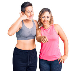 Couple of women wearing sportswear smiling doing talking on the telephone gesture and pointing to...