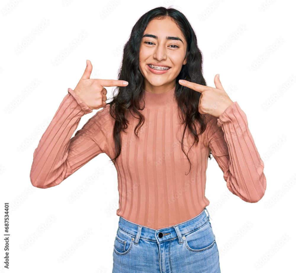 Wall mural Hispanic teenager girl with dental braces wearing casual clothes smiling cheerful showing and pointing with fingers teeth and mouth. dental health concept.