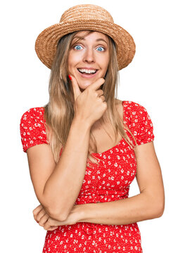 Beautiful caucasian woman wearing summer hat looking confident at the camera smiling with crossed arms and hand raised on chin. thinking positive.
