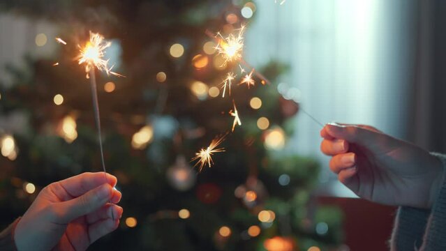 Closeup of hands of couple holding two stick of holiday sparkler burning on Christmas tree bokeh background tree at home, Celebrate Christmas and New Year.