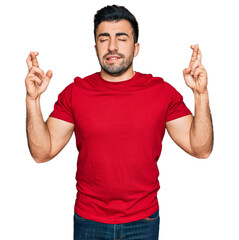 Hispanic man with beard wearing casual red t shirt gesturing finger crossed smiling with hope and eyes closed. luck and superstitious concept.