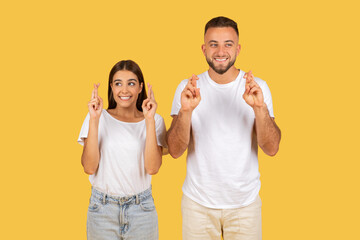 Happy millennial european wife and husband in white t-shirts with crossed fingers hoping or wishing
