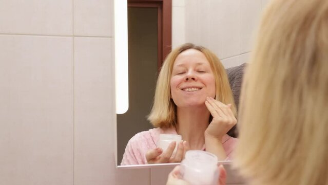 Collagen facial cream, woman smearing her face with a moisturizing product in the bathroom in front of the mirror, Cosmetics for skincare and makeup, skin treatment