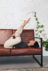 Attractive brunette woman performs a variation of the Supta Ekapada Agni Stambhasana exercise, practicing yoga while lying on the sofa in casual clothes