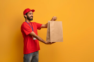 Cheerful delivery man in red uniform and cap extending brown paper bag