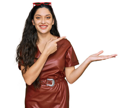 Brunette young woman wearing dress and sunglasses amazed and smiling to the camera while presenting with hand and pointing with finger.